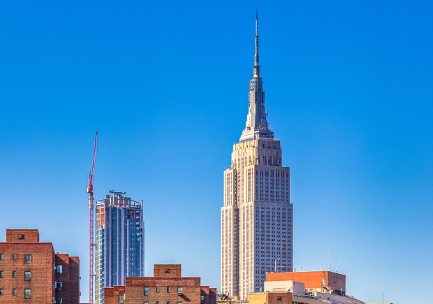 A photo of a blue sky and New York City buildings with a river in the foreground, Peter Cooper Village buildings and the Empire State building in the midground, and a skyscraper under construction in the background